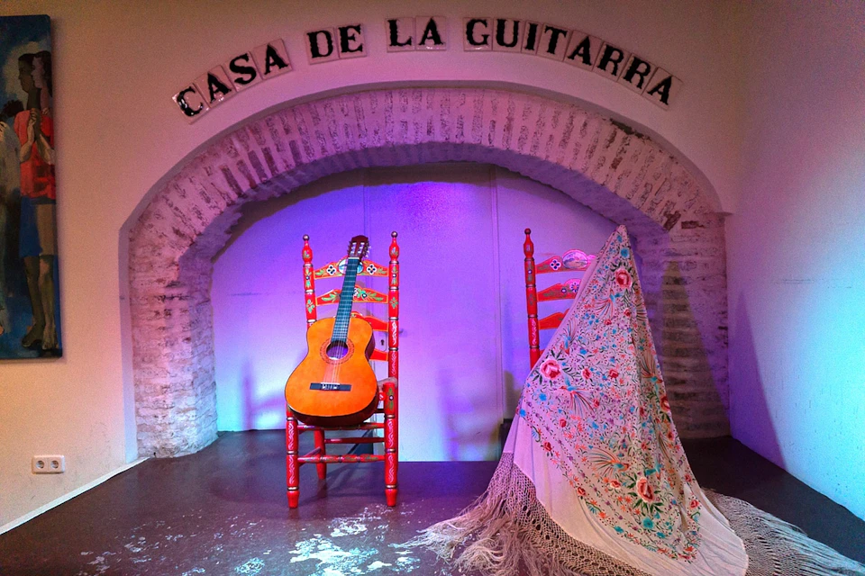 A stage with a sign reading "Casa de la Guitarra," featuring a guitar on an ornate red chair and another chair draped with a colorful shawl.