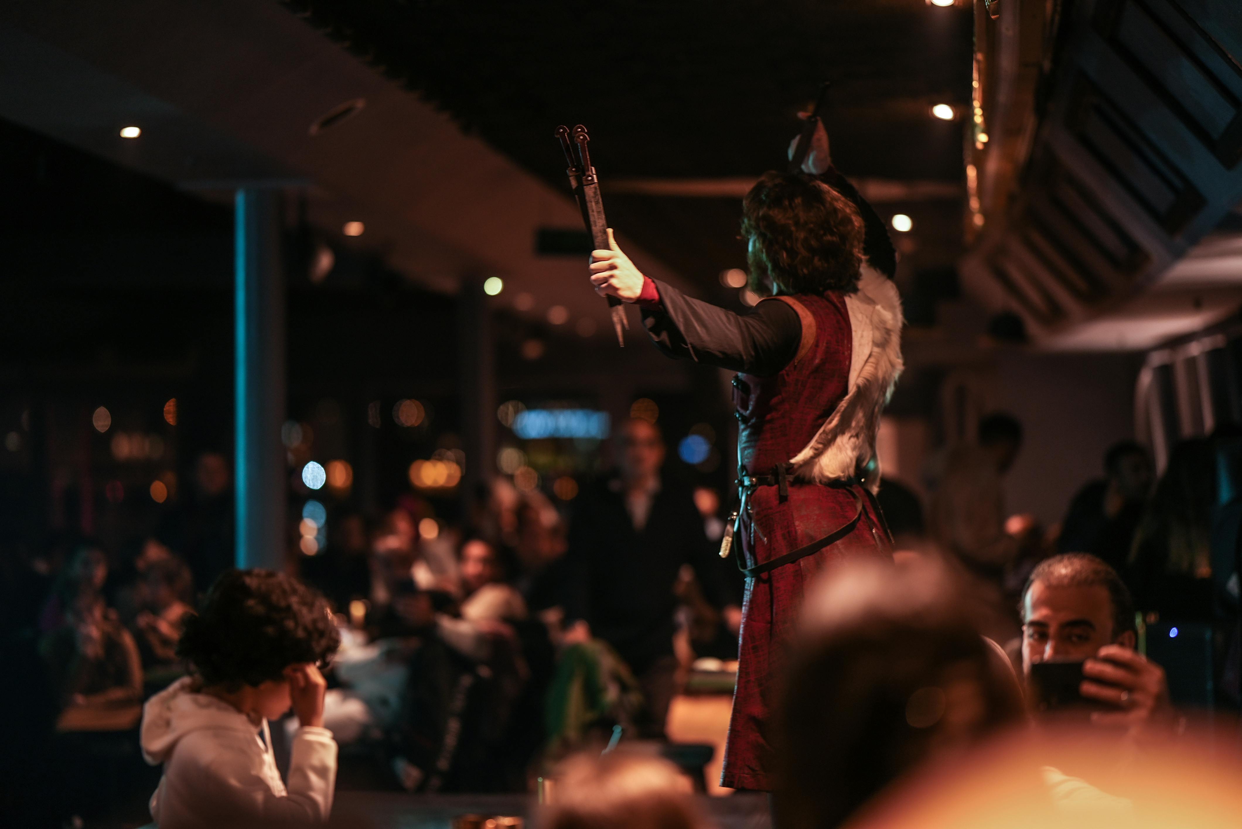 歴史的な服装をした人物が、薄暗い部屋で腕を上げてジェスチャーをしており、観客が見ています。