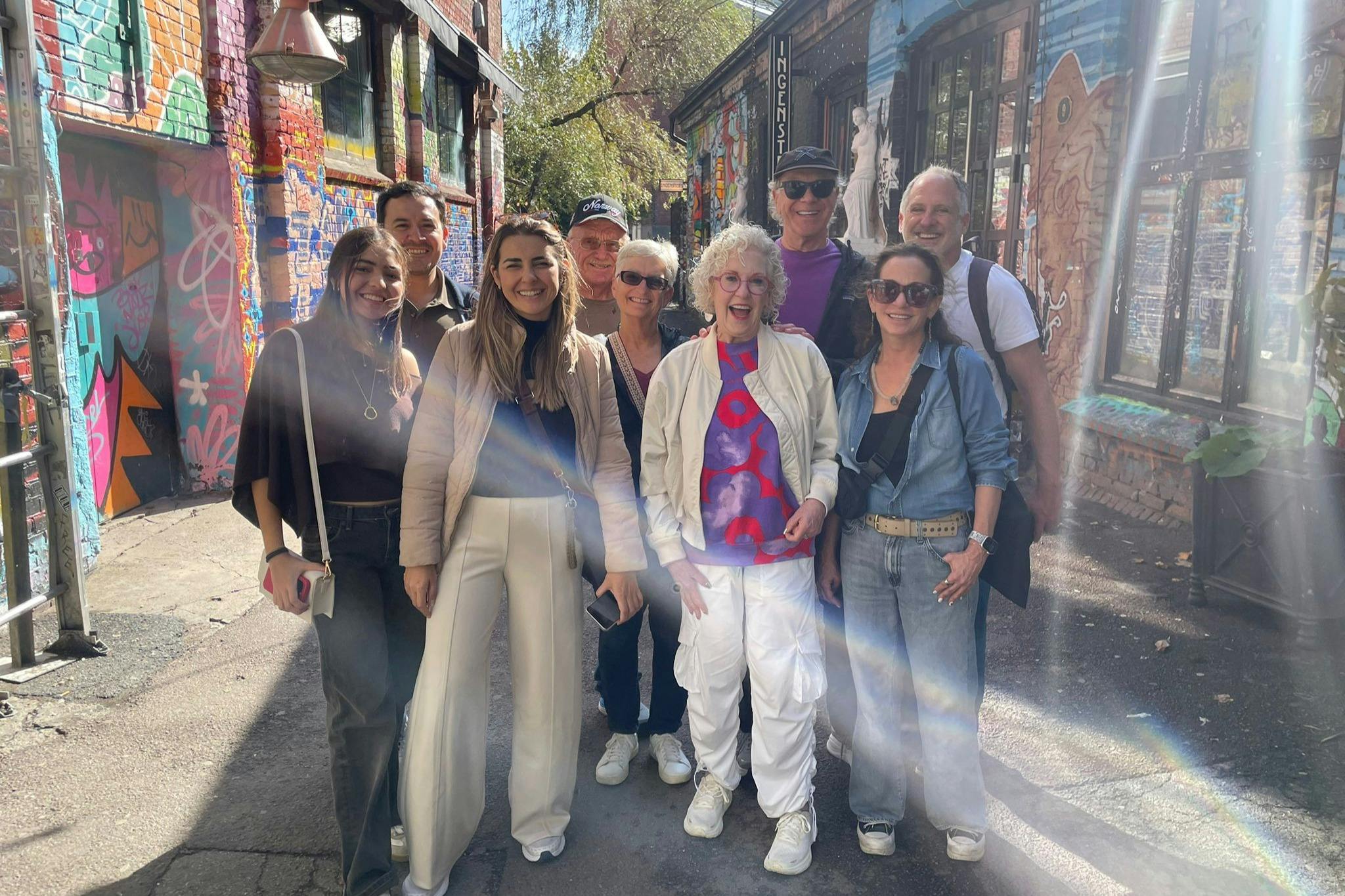 A group of nine people smiling and posing in a colorful alley with vibrant graffiti-covered walls.