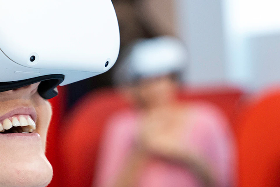 A person smiling while wearing a virtual reality headset, seated indoors with another person blurred in the background.