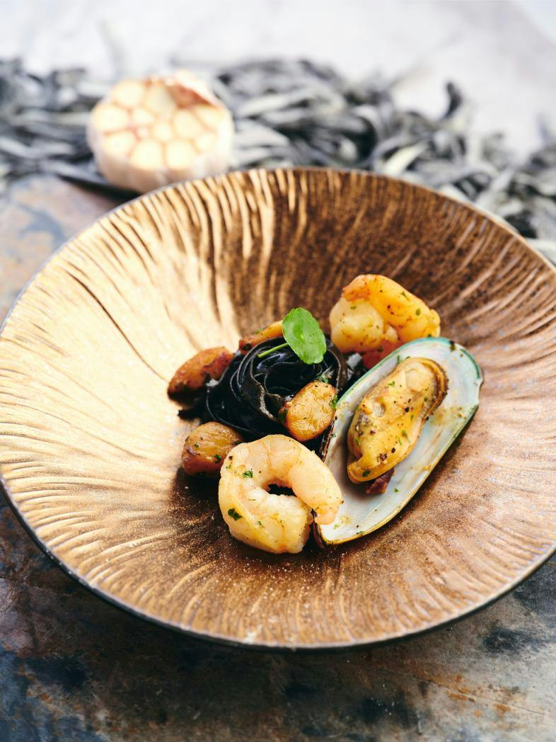 A dish of black pasta with shrimp, mussel, and garnishes served in a textured golden bowl on a rustic surface.