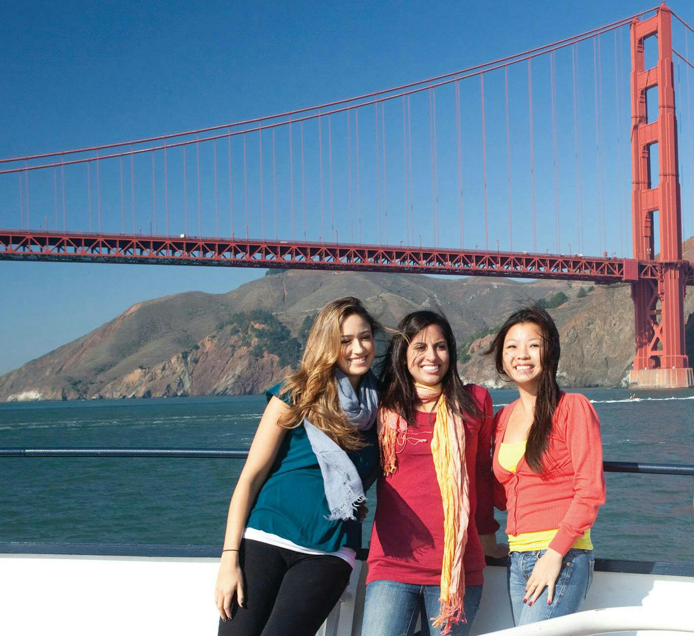 三个女人在金门大桥前微笑，背景是晴朗的蓝天和丘陵景观。