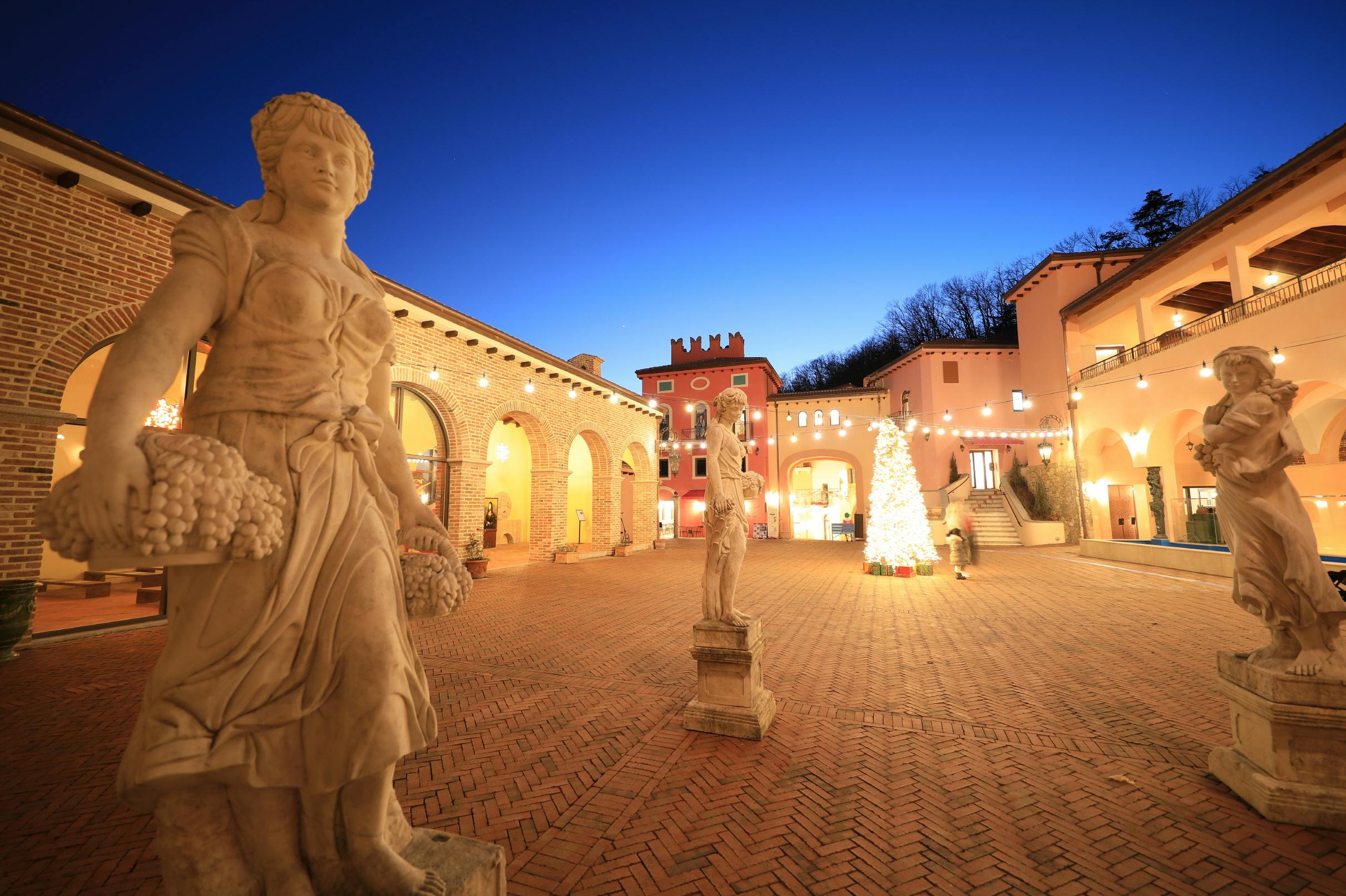 Outdoor courtyard with brick buildings, statues holding baskets, and warm lighting against a twilight sky.