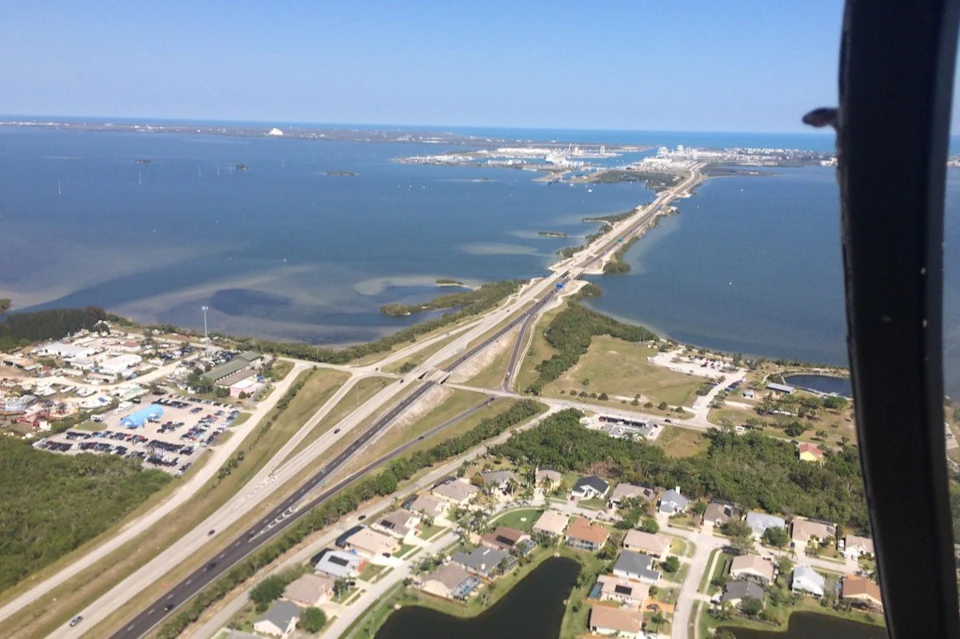 Aerial view of a coastal highway bridge connecting land over water, with nearby residential and industrial areas under a clear sky.