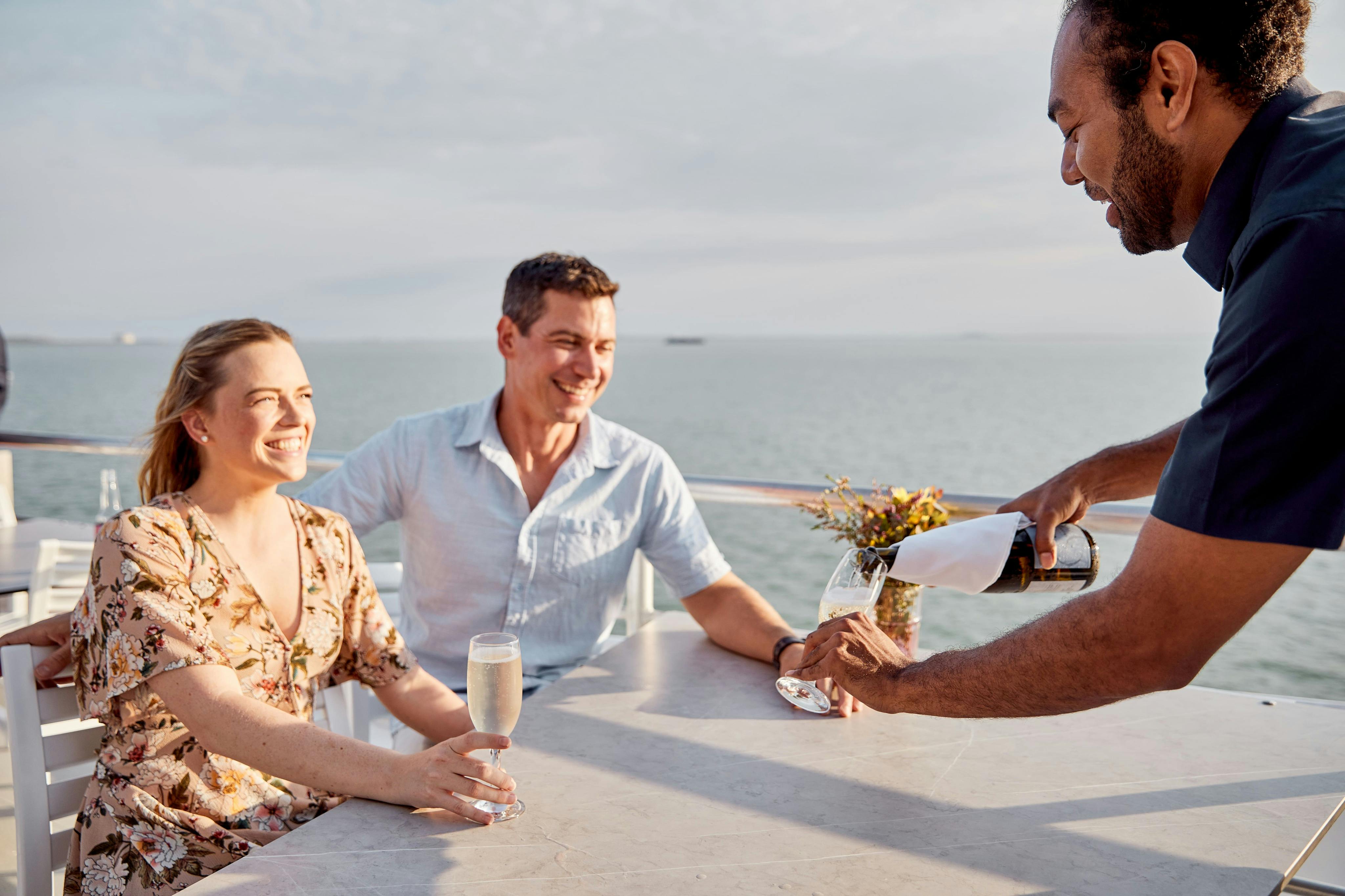 ボートの屋外テーブルに座っているカップルは、サーバーがシャンパンをグラスに注ぎ、海の景色を背景に微笑んでいます。