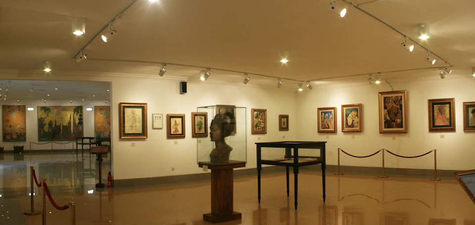 Art gallery interior with framed paintings on the walls, a bust in a glass case on a wooden pedestal, and an empty display table.