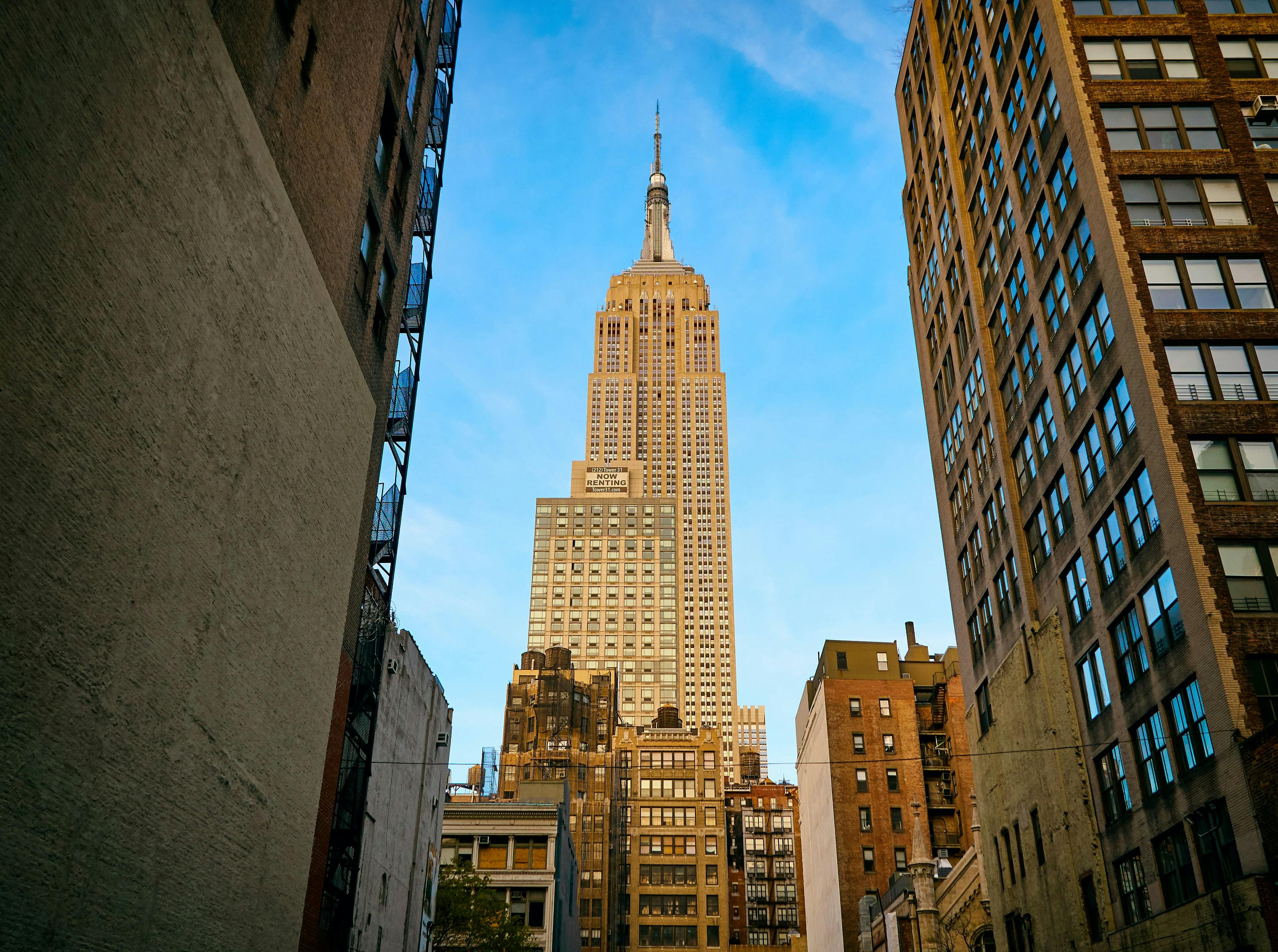 Άποψη του Empire State Building από έναν δρόμο που πλαισιώνεται από ψηλά κτίρια, κάτω από έναν καταγάλανο ουρανό.