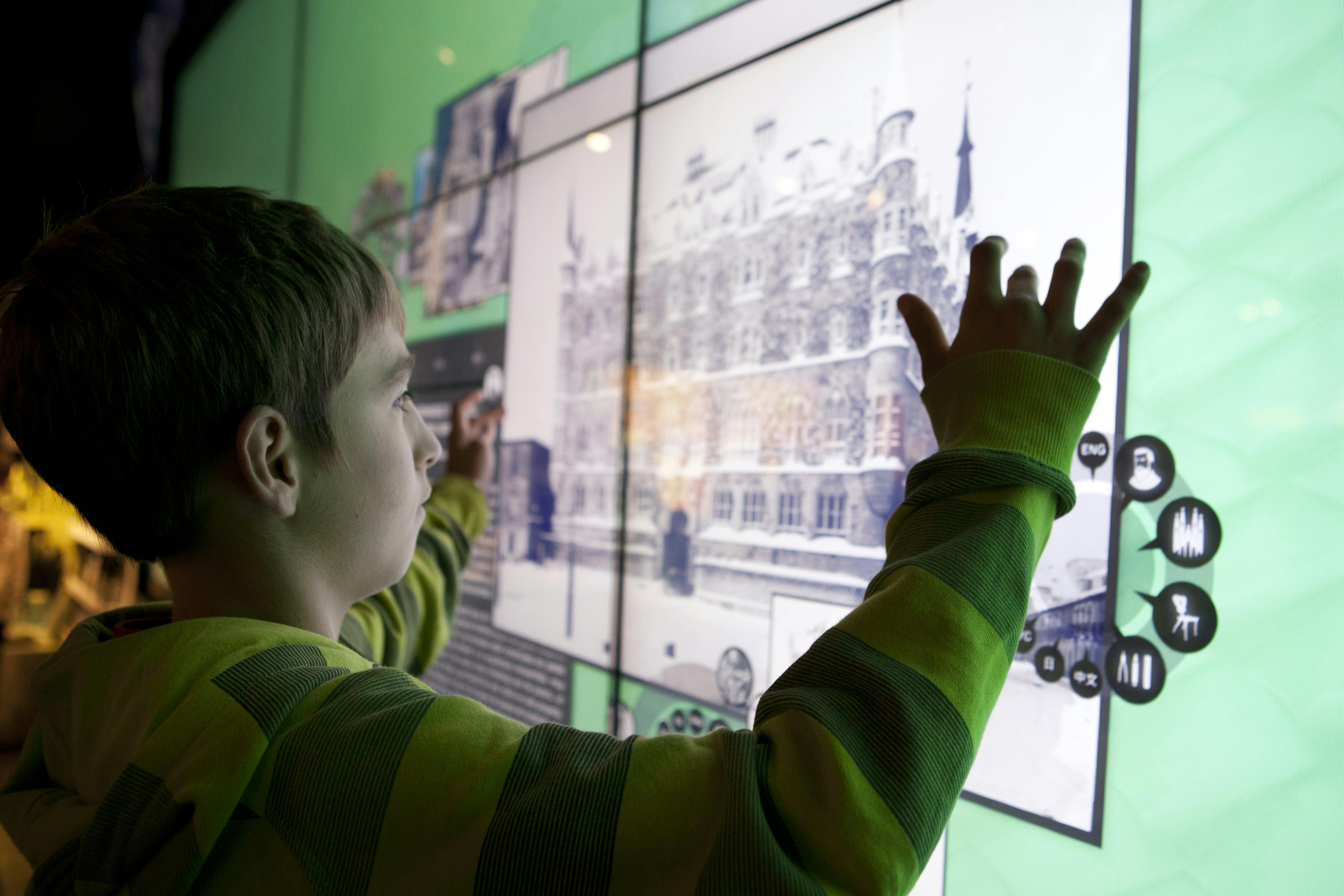 緑色の縞模様のパーカーを着た人物が、建物の歴史的な画像を表示する大きなタッチスクリーンと対話しています。