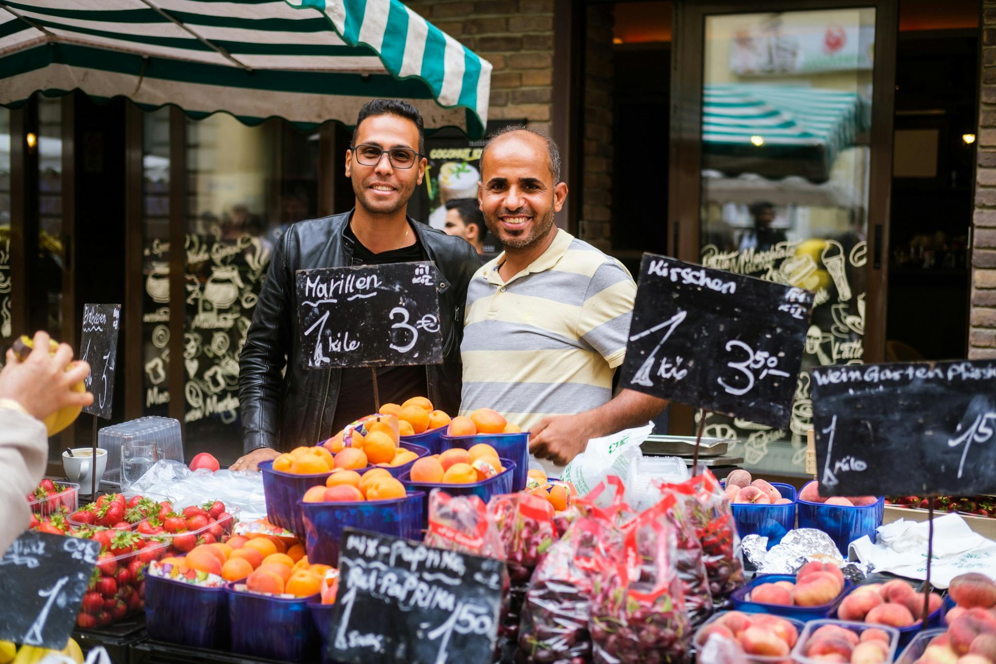 Dva muži stojí za stánkem s ovocem na venkovním trhu. V tabulce jsou jahody, broskve a cedule s cenami.