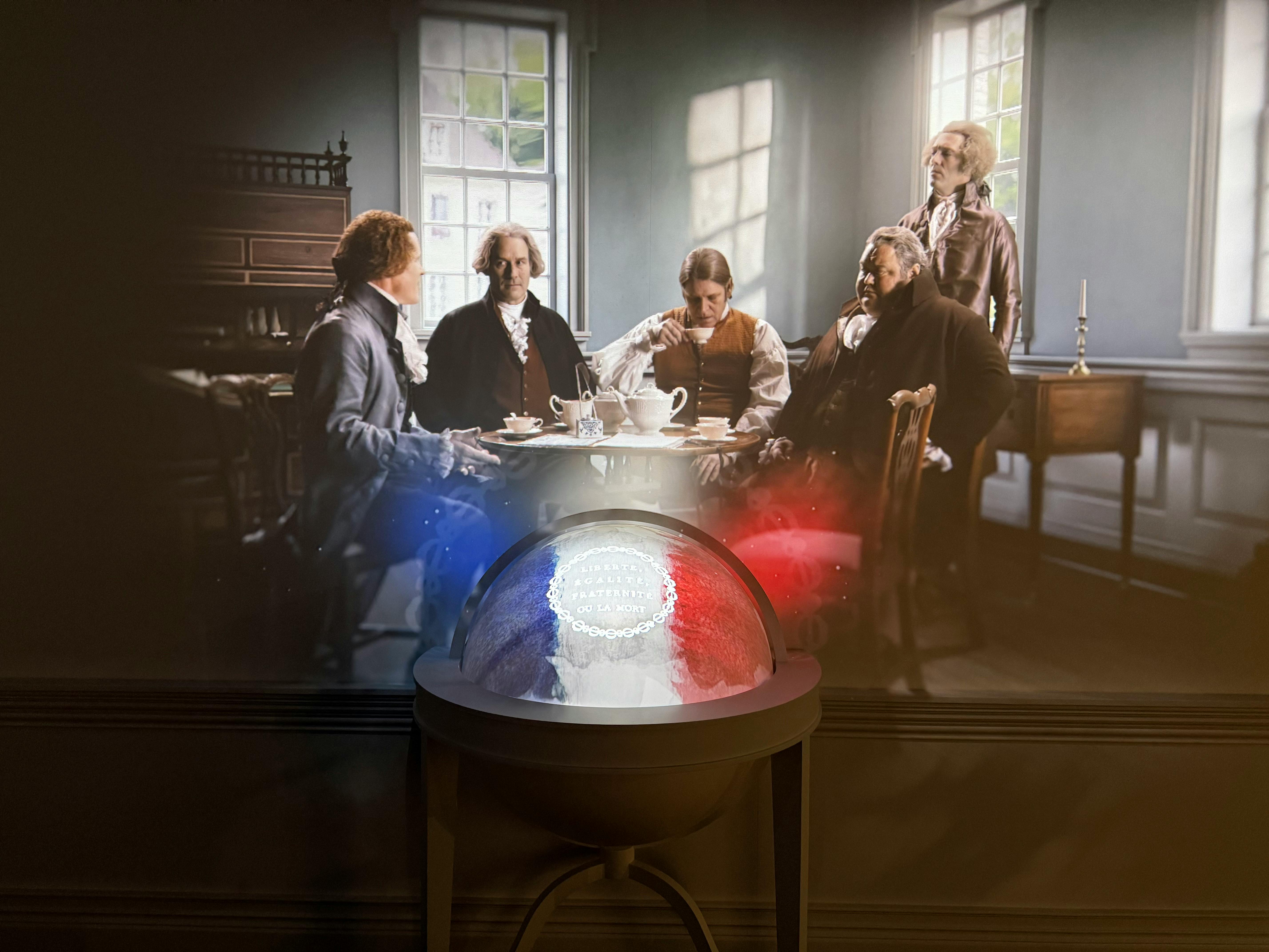 A group of five figures in historical attire sit around a table in a room, with a glowing globe-like object in the foreground.