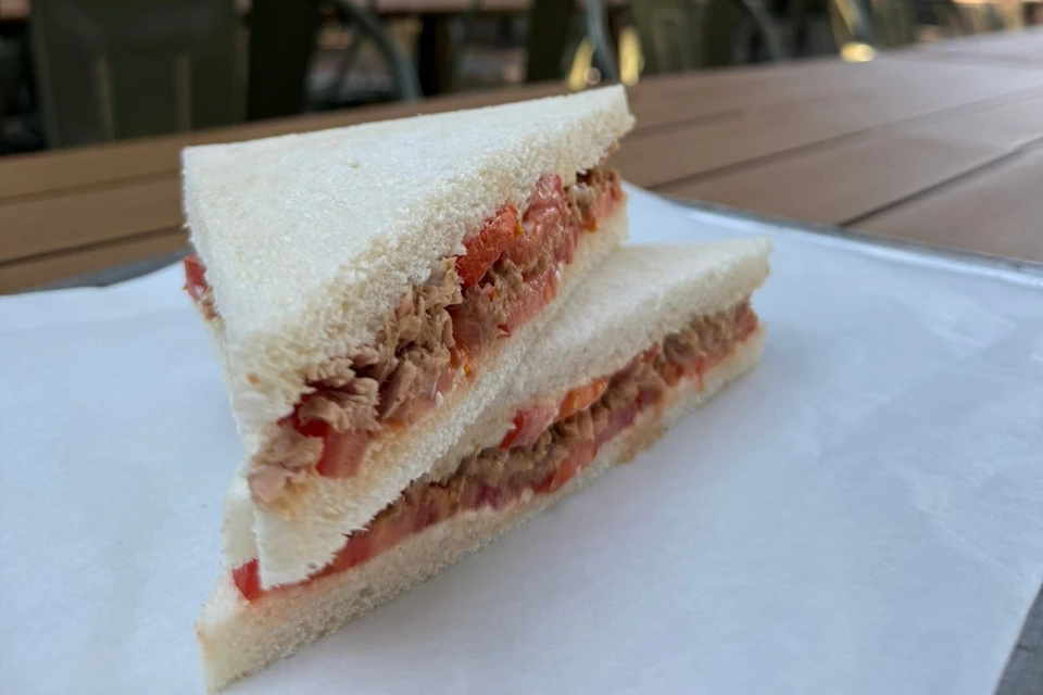 Close-up of a sandwich with white bread, tuna, and tomato slices, placed on a piece of parchment paper on a wooden table.