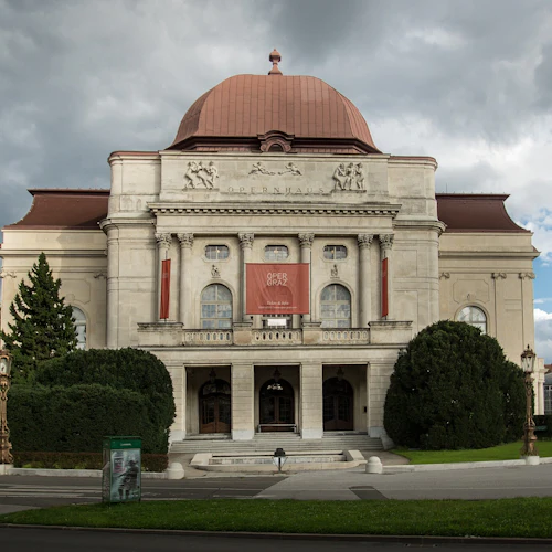 En utsmyckad byggnad med klassisk arkitektur märkt "Opernhaus", med ett välvt tak, pelare och en välskött gräsmatta.