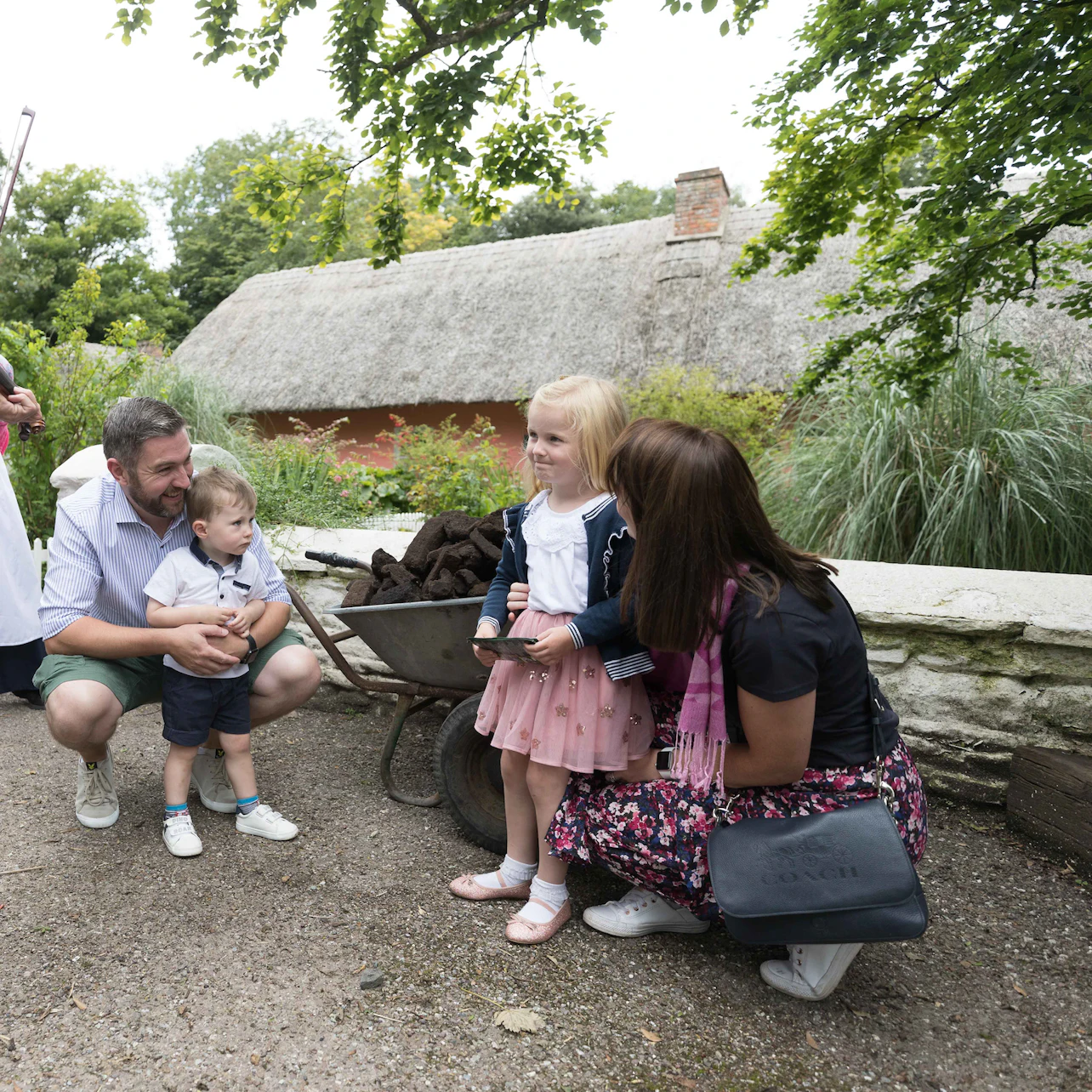 Two adults and two children are observed near a wheelbarrow filled with peat, outside of a thatched-roof building.