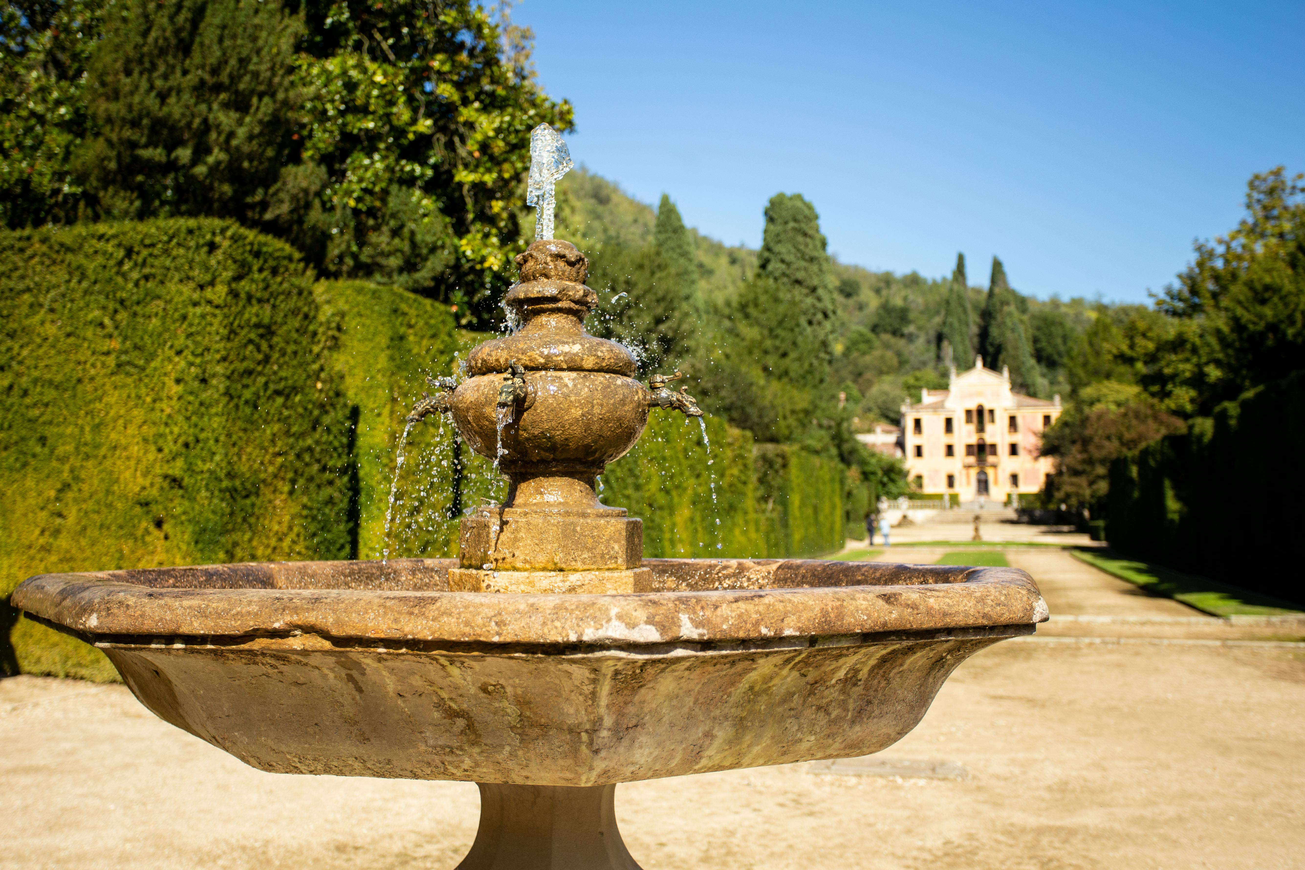 Una fontana in pietra con getti d'acqua in un giardino, affiancata da alte siepi e alberi, con un grande edificio sullo sfondo.