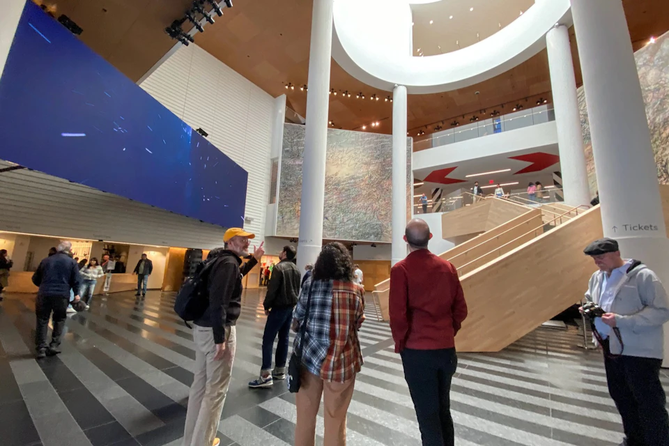 People standing in a modern, multi-story atrium with a large wall display, wooden stairs, and a mix of natural and artificial lighting.