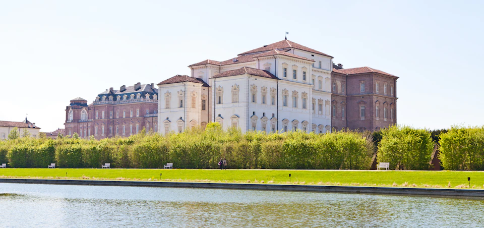 Large, historic white building with ornate windows and red roofs, bordered by greenery and a body of water in the foreground.