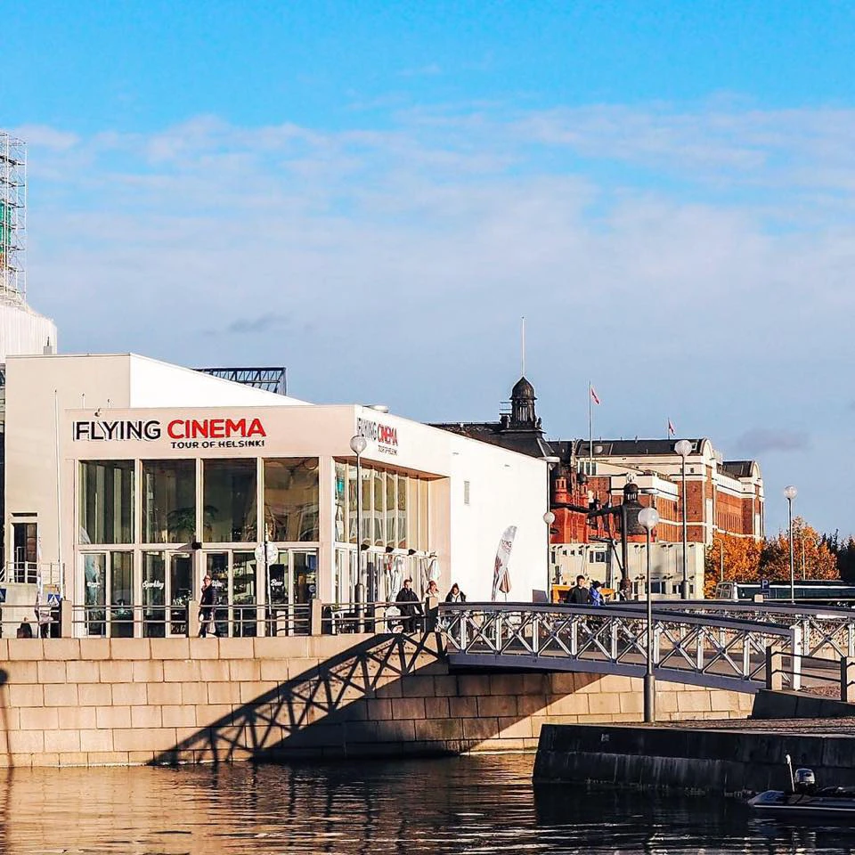 Building with signage "Flying Cinema Tour of Helsinki" next to a waterfront and bridge, with people walking nearby on a sunny day.