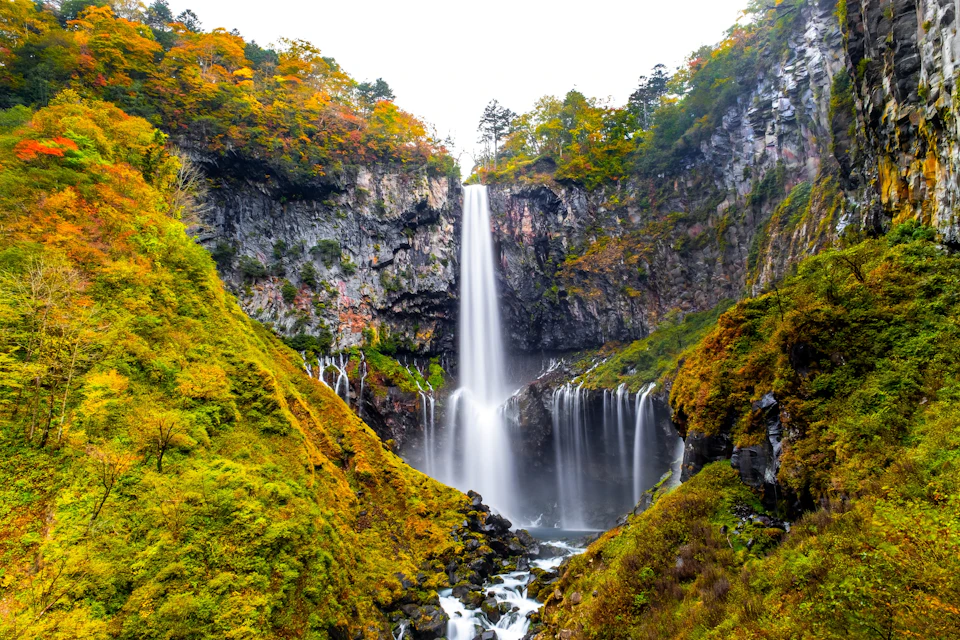 A tall waterfall cascades into a rocky basin, surrounded by colorful autumn foliage and steep cliffs.