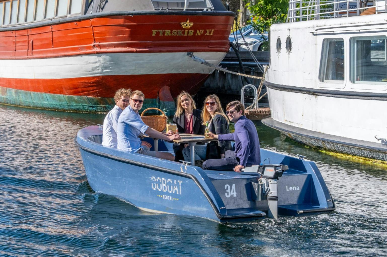 Five people smiling and sitting around a table on a blue boat named Lena, with larger boats docked in the background.