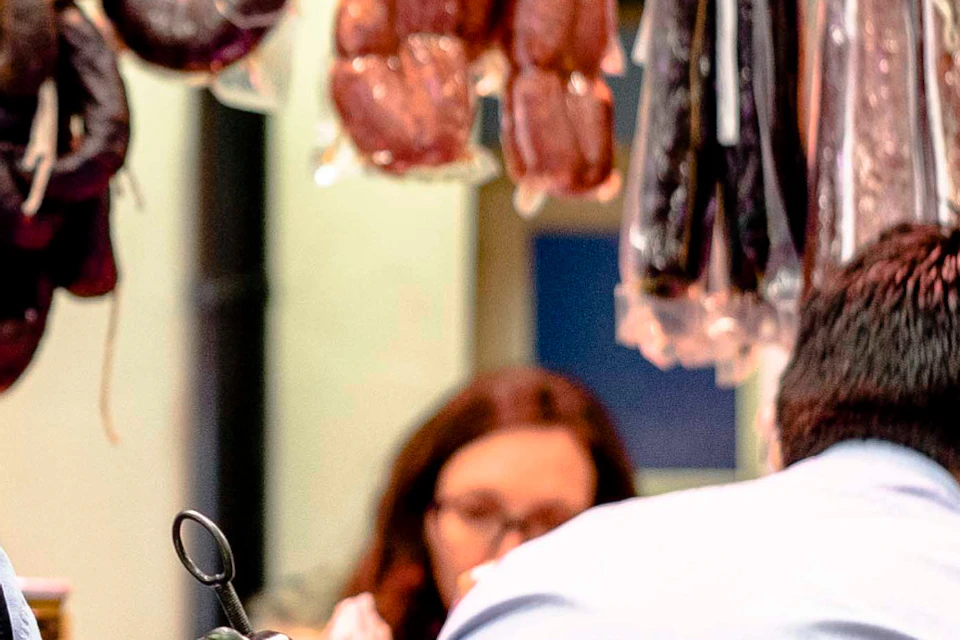 Man in a deli holding a large cured ham leg. Assorted meats hang behind him, with glassware and jars on the counter.