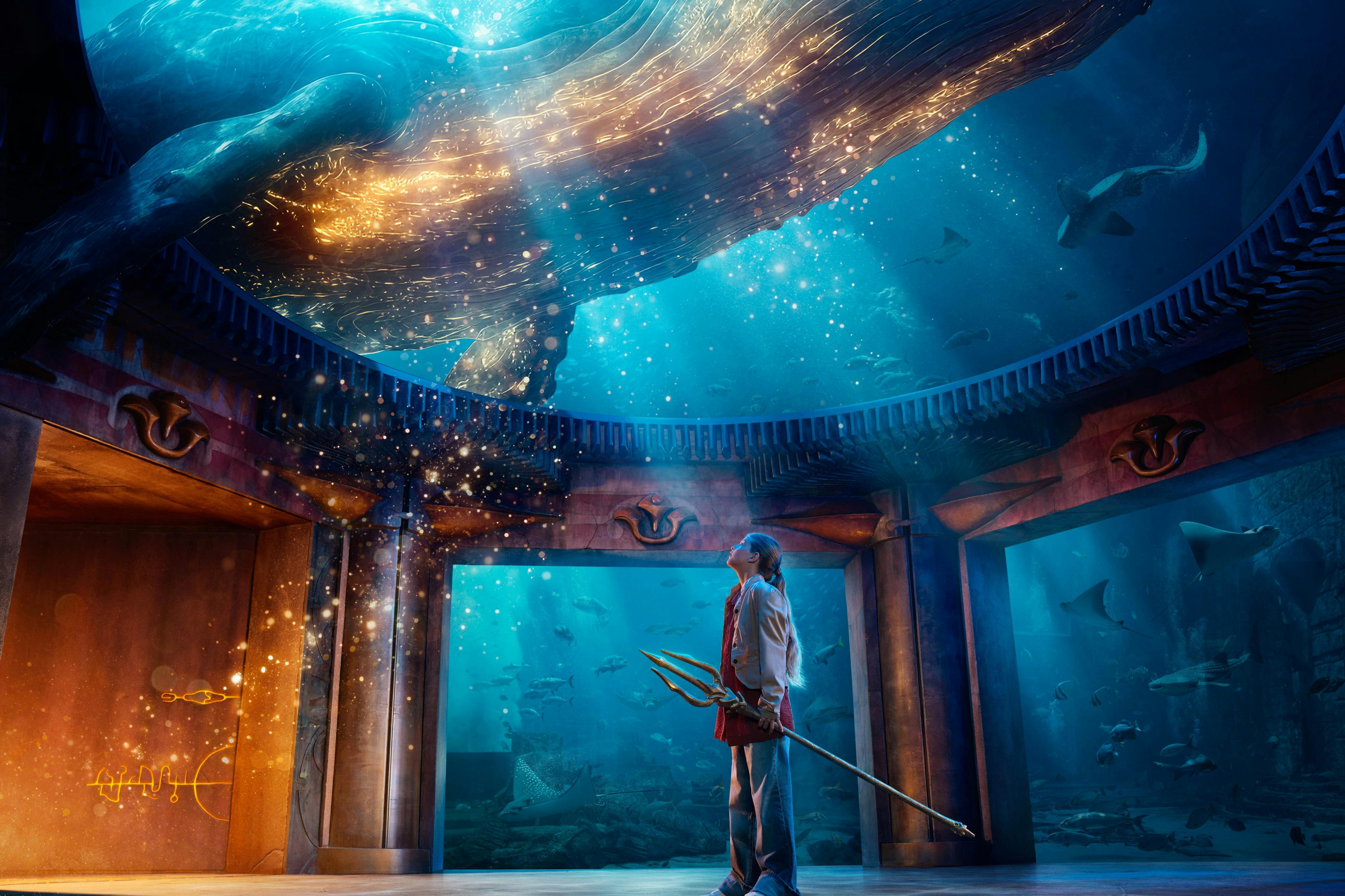 Eine Person mit einem Dreizack steht in einer Unterwasserhalle und blickt auf einen leuchtenden Wal, der über ihr schwimmt.