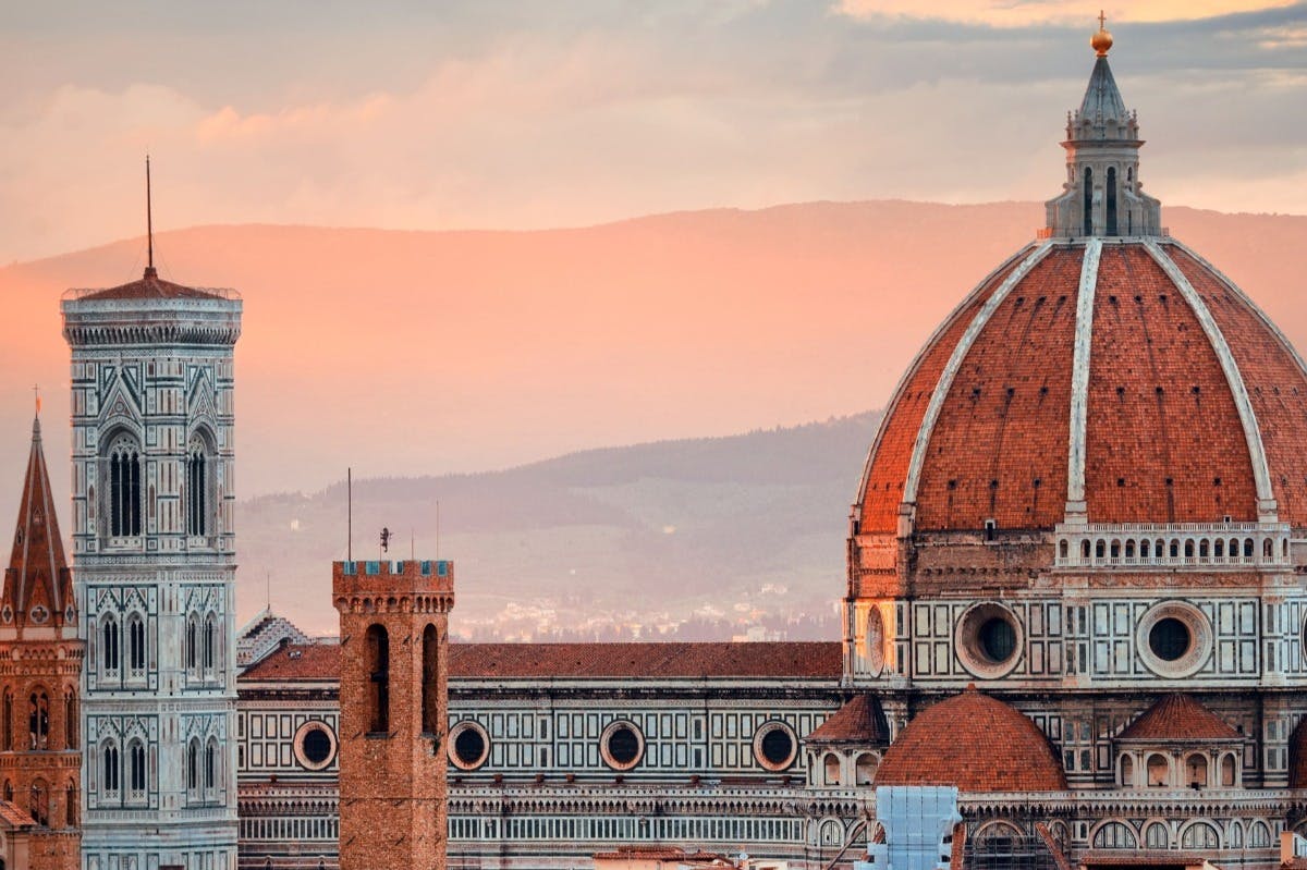 Vue de la cathédrale de Florence avec son dôme caractéristique, une tour en brique et des montagnes en arrière-plan sous un ciel rosé.