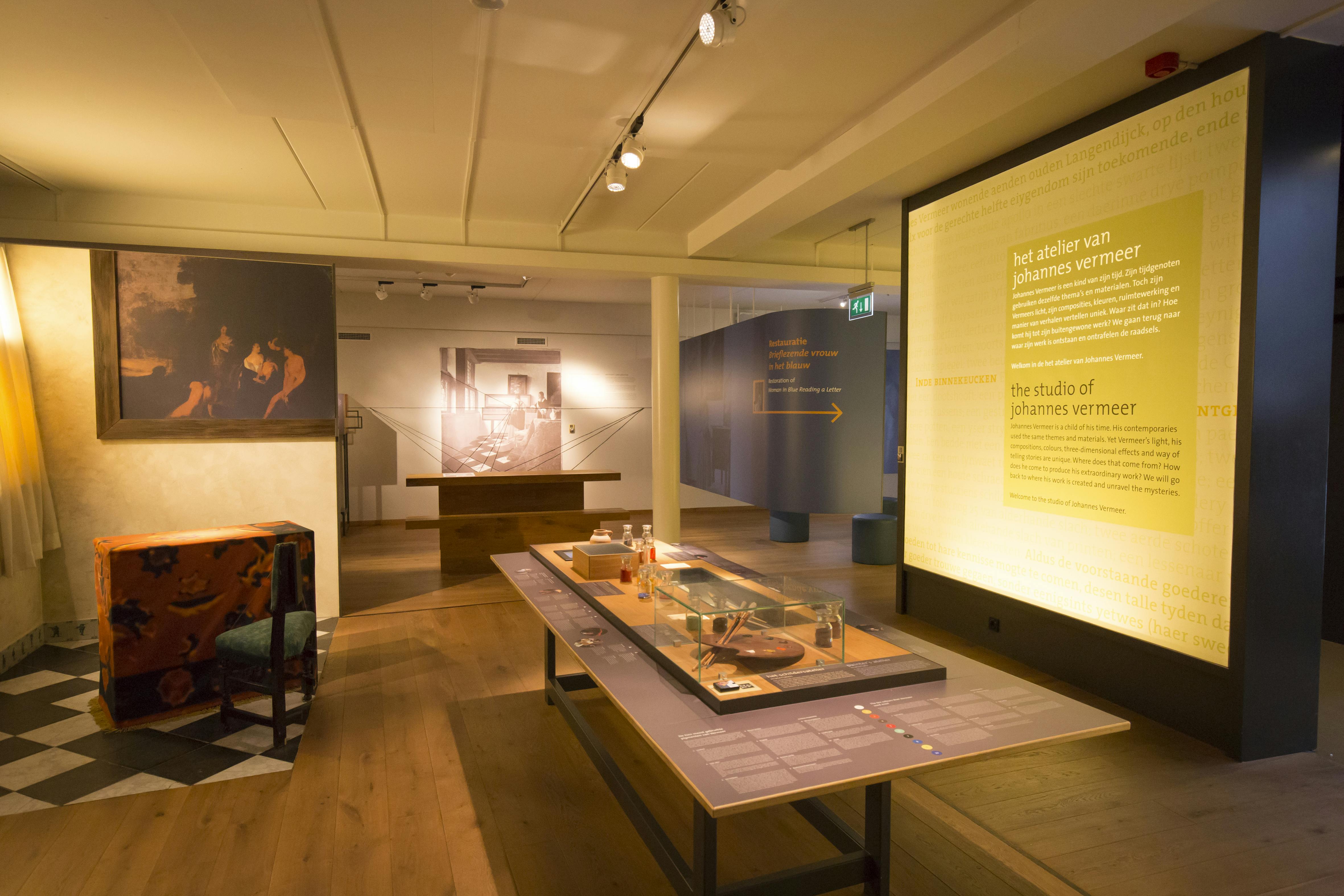 Uma sala de exposições do museu com exposições informativas sobre Johannes Vermeer, incluindo painéis de texto, fotografias e uma mesa central.