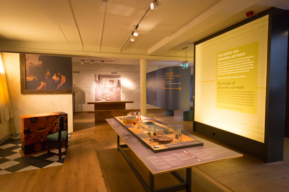 A museum exhibit room with informational displays about Johannes Vermeer, including text panels, photos, and a central table.