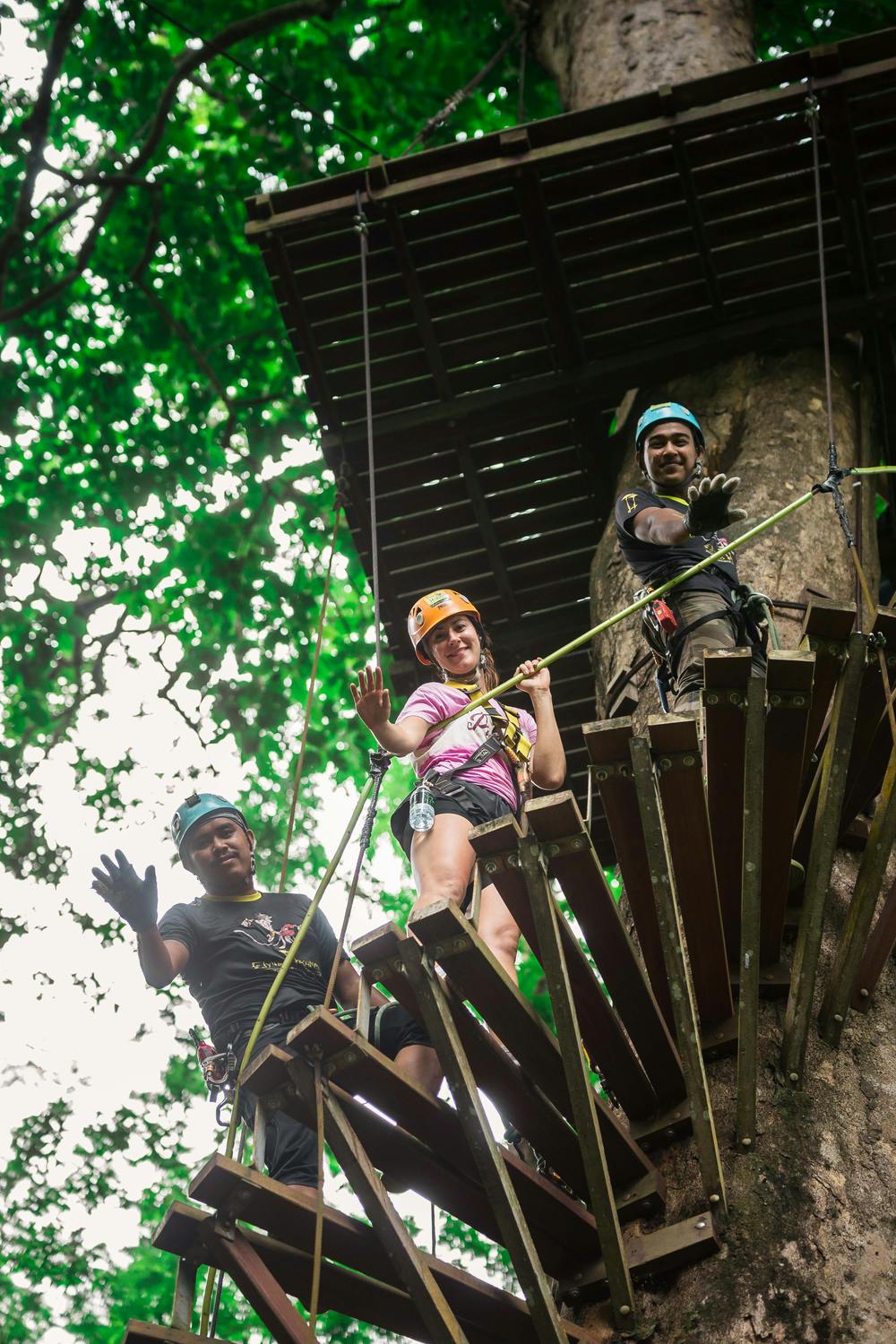 三个人戴着头盔和马具，站在树丛中高高的木制平台上，对着镜头挥手。