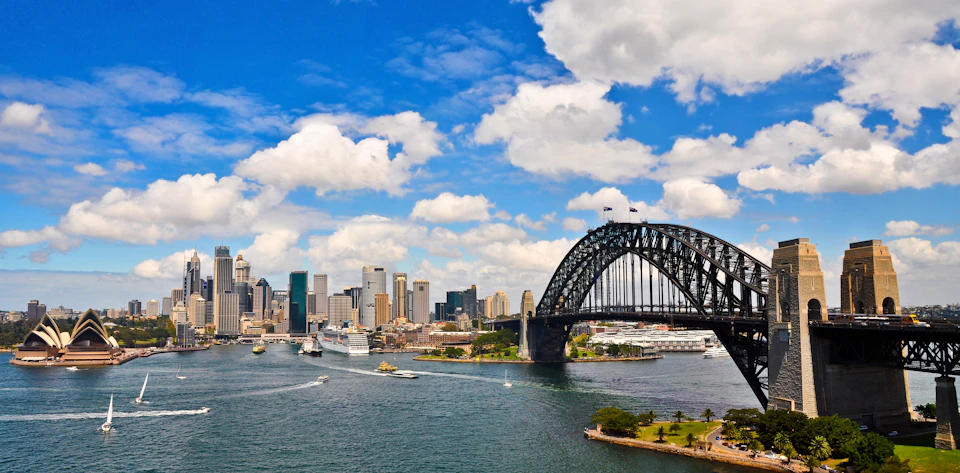 City skyline with tall buildings and a bridge over water under a blue sky with clouds. Boats and cruisers are on the water.