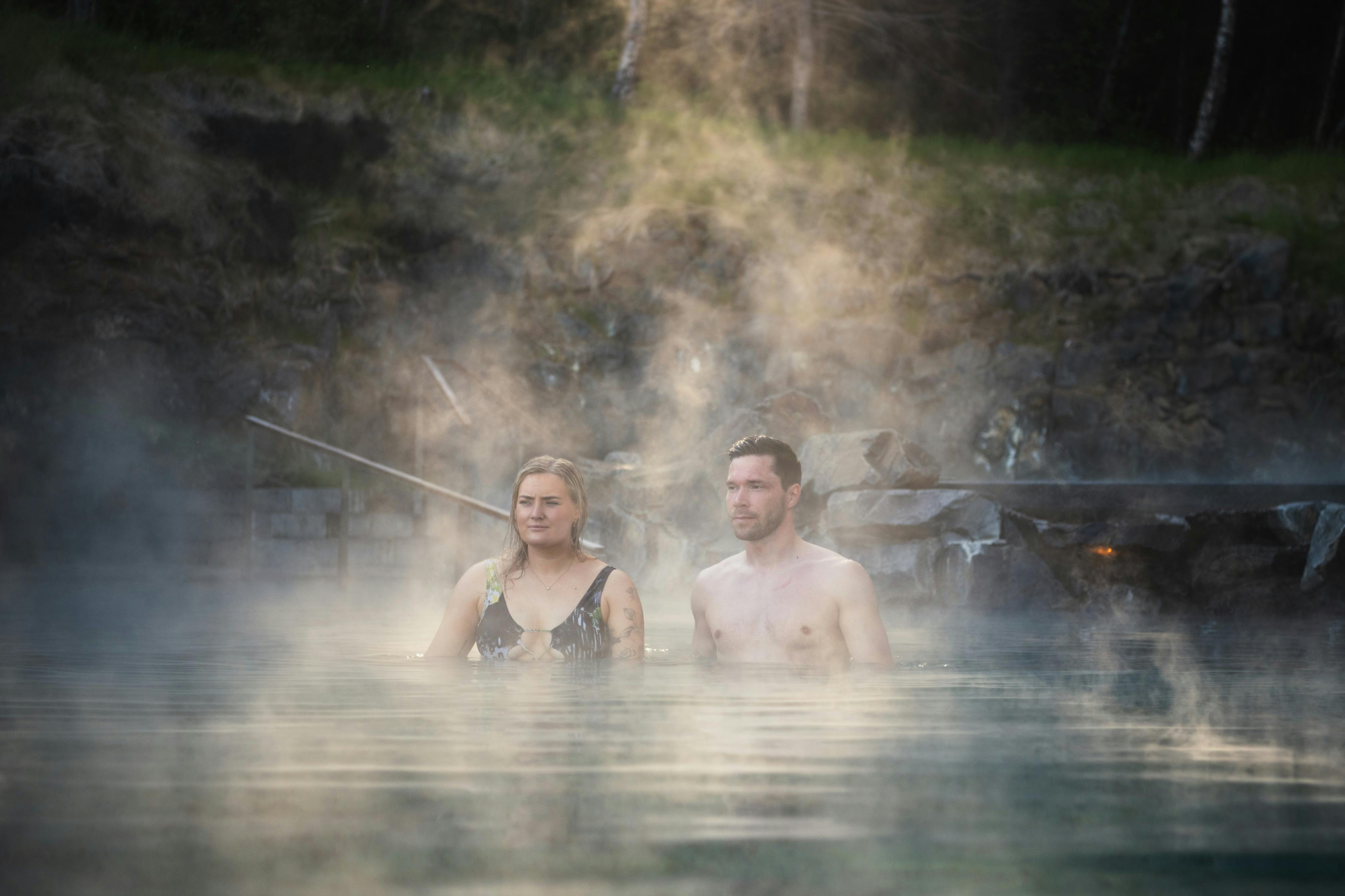 Två personer står i en ångande naturlig varm källa omgiven av stenig terräng och grönska.