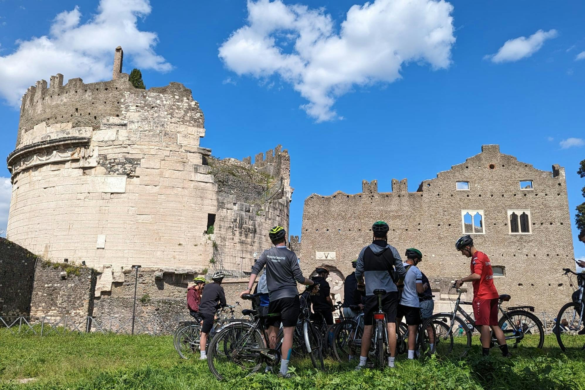Grupa rowerzystów stojących ze swoimi rowerami w pobliżu starożytnych ruin w słoneczny dzień z błękitnym niebem i chmurami.