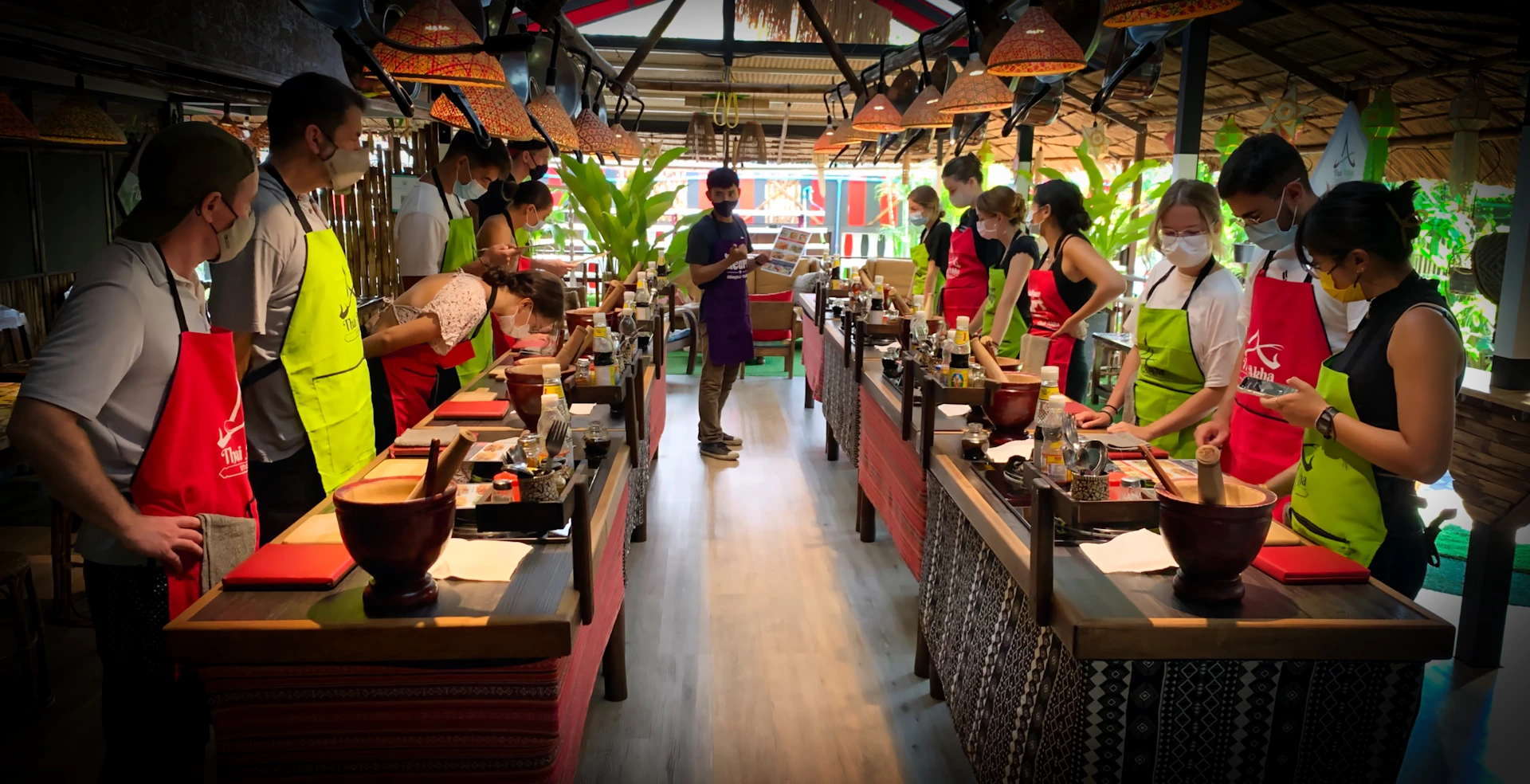A group of people wearing aprons participate in a cooking class in a tropical-themed setting with wooden tables and tools.