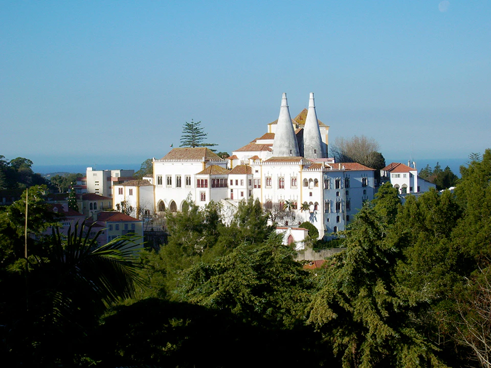 Large white palace with two distinctive conical chimneys, surrounded by greenery and set against a clear blue sky.