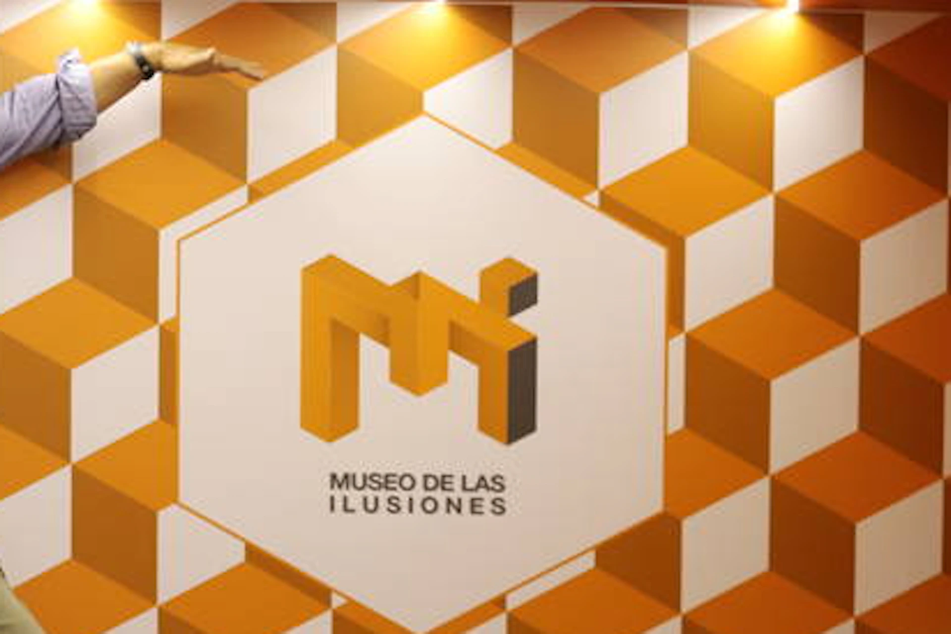 Two people pose in a room with an optical illusion, making one appear significantly larger than the other. Walls have an orange-and-white 3D cube pattern.