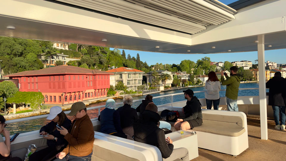 People seated on a boat deck with a view of a waterfront featuring colorful buildings and greenery in the background.