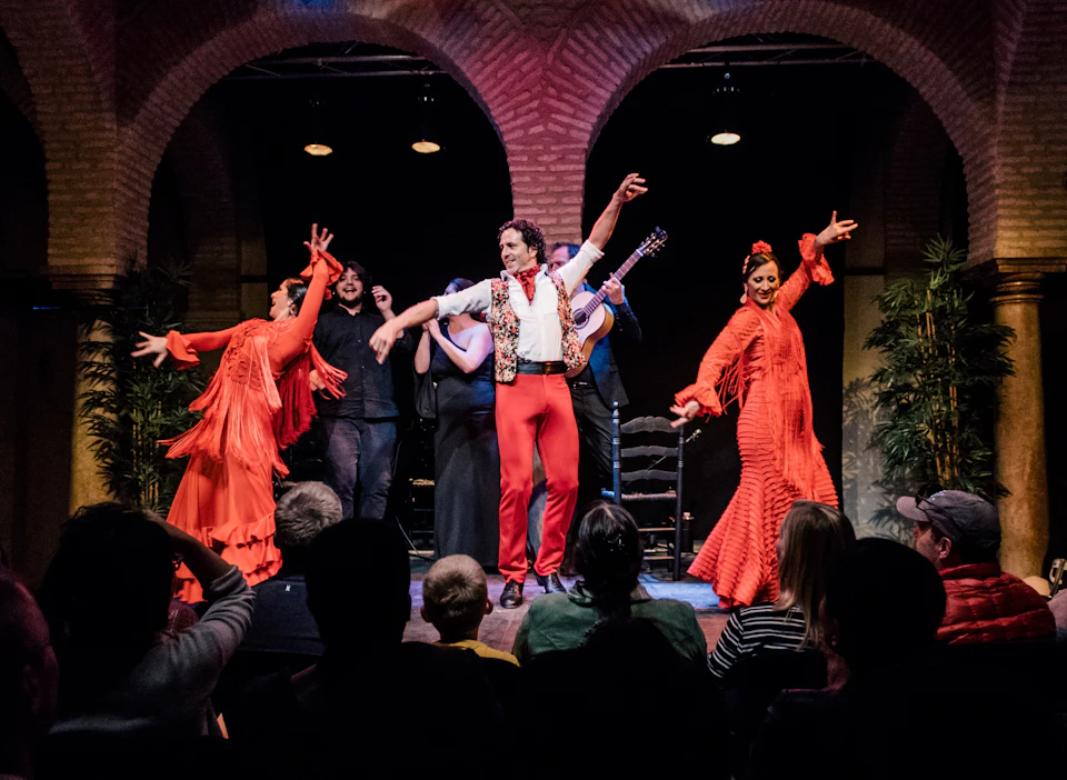 Flamenco dancers in vibrant red costumes perform on a stage, accompanied by musicians, as an audience watches.