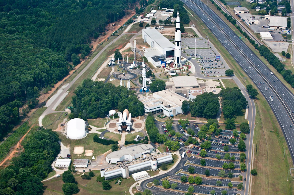 Aerial view of a space center with rockets, space shuttle replicas, buildings, parking lots, and surrounding trees.