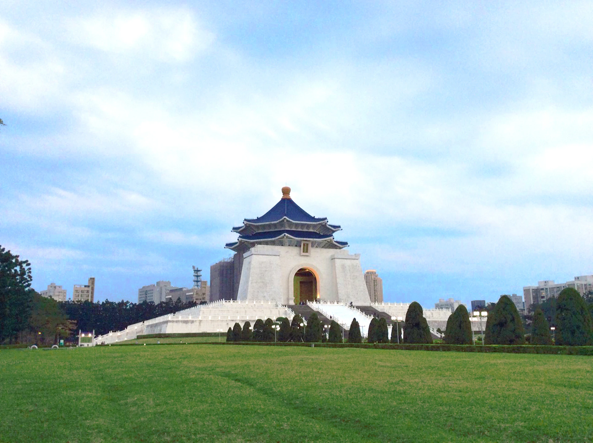 A grand white building with a blue roof and ornate architecture stands in a large green park under a partly cloudy sky.