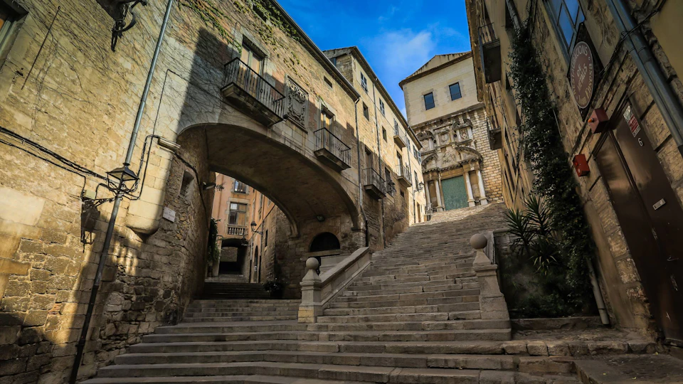 Stone staircase leading up between historic buildings under a blue sky, with an archway and ornate facade visible.