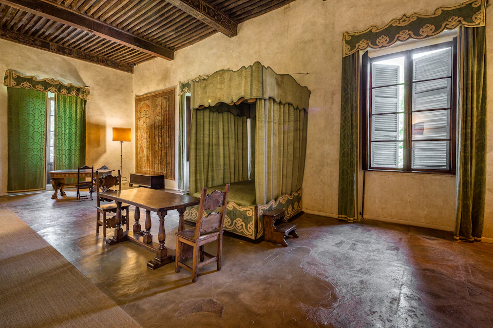 A historic room features a canopy bed, wooden furniture, a lit floor lamp, a tapestry, and an open window with shutters.