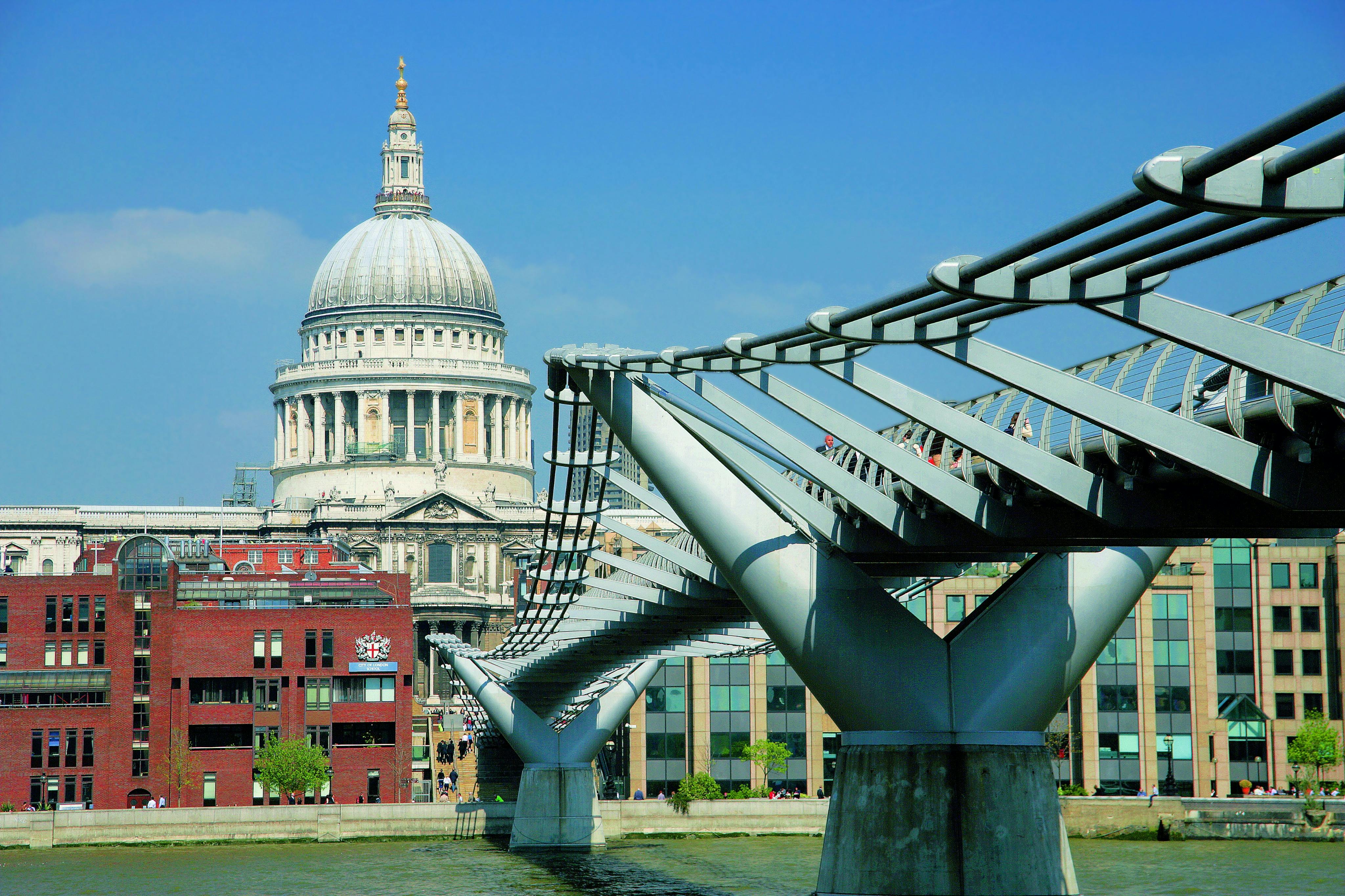 A modern steel pedestrian bridge spans a river with a historic domed building and cityscape in the background under a clear sky.
