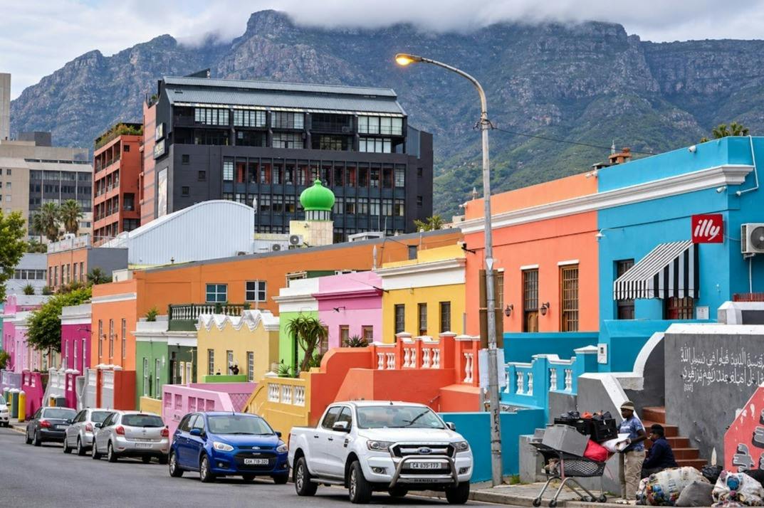 Colorful houses on a street with parked cars, a mountain in the background, and a cloudy sky.
