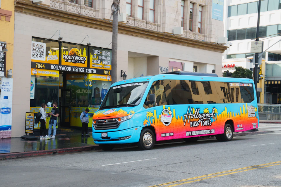 A bright blue Hollywood tour bus parked in front of a visitor center building with a sign reading 'Hollywood Visitor Center.'