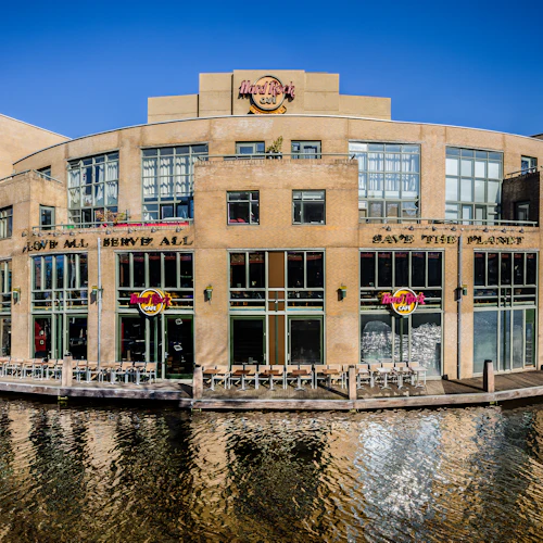 Hard Rock Cafe-byggnad i tre våningar med sittplatser på uteplatsen vid vattnet, synlig skyltning och en klarblå himmel.