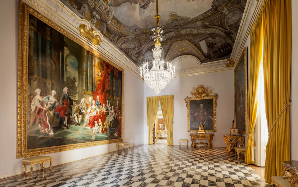 Luxurious room with a large chandelier, baroque paintings on the walls, ornate gold furnishings, and checkered marble flooring.