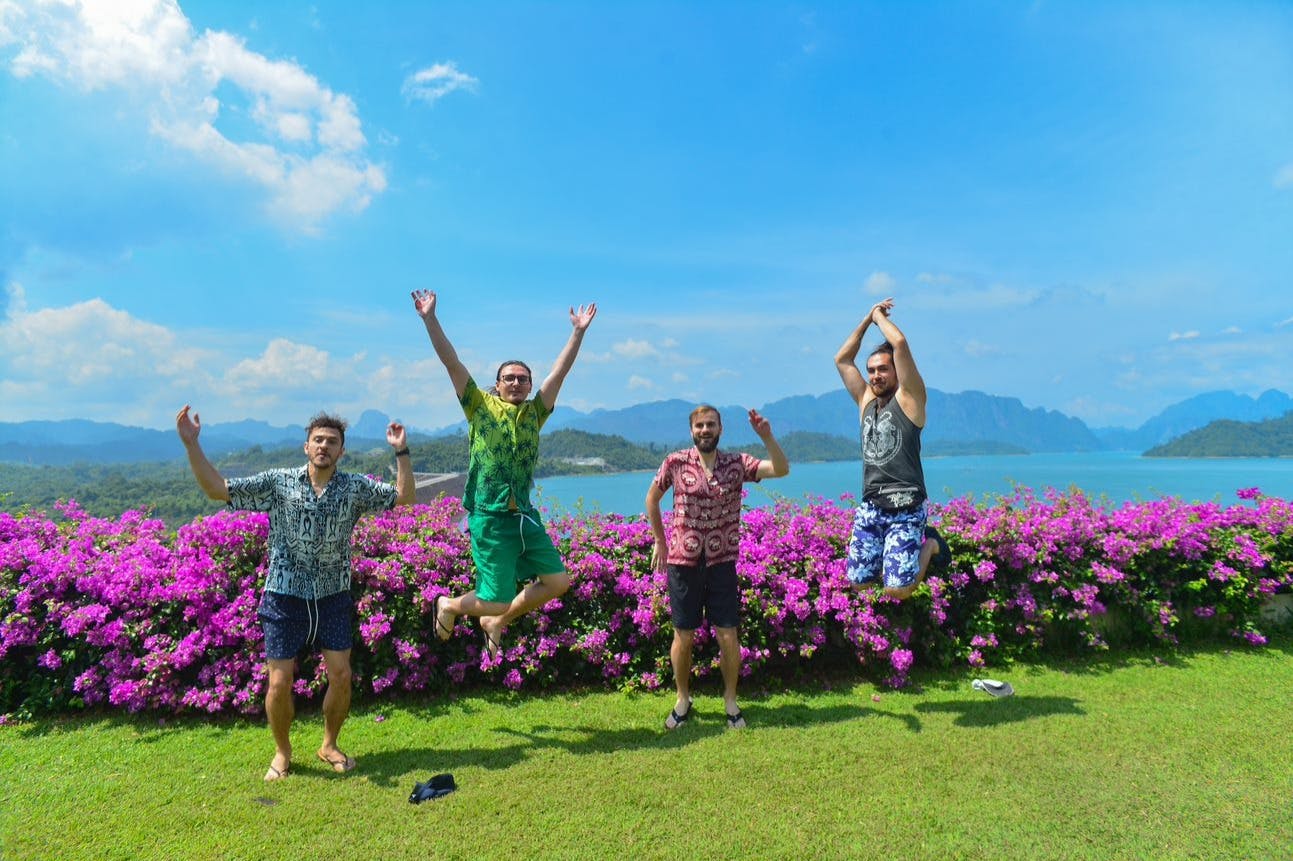 四名身穿五颜六色衬衫的男子在盛开的粉红色花朵前摆姿势，两人跳跃，山湖景色优美。
