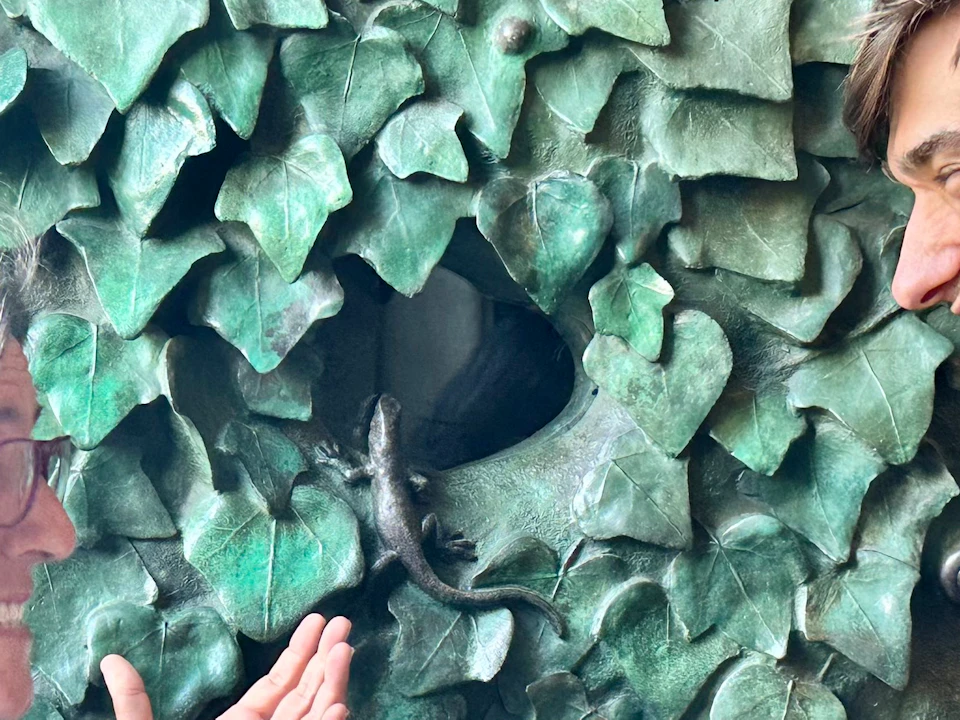 Two people smiling at each other in front of a green, leafy textured background, one with glasses and both gesturing with their hands.