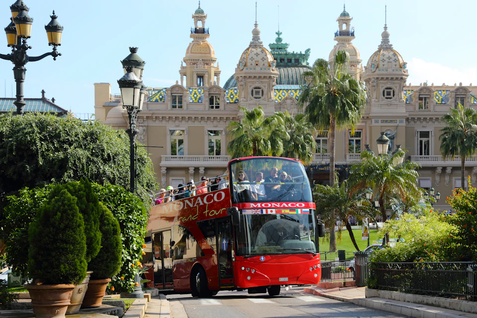 A red double-decker tour bus labeled "Monaco" drives past ornate buildings and palm trees under a clear sky.