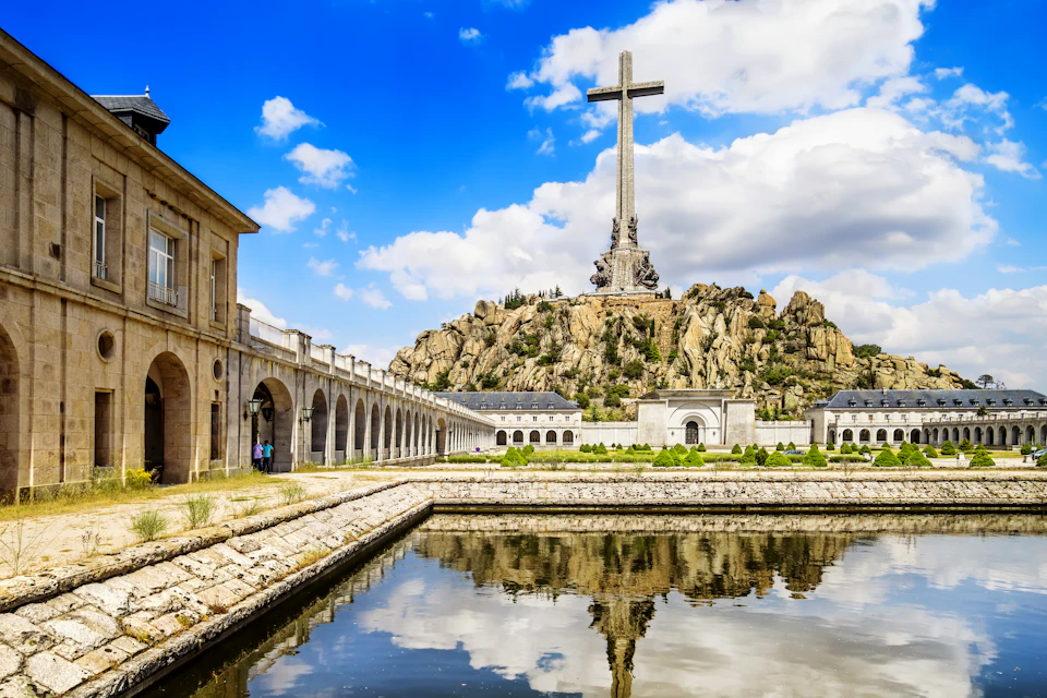 Massive cross on a rocky hill with an arched colonnade to the left and a reflective pool in the foreground under a blue sky.